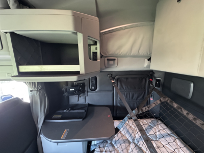 Kenworth T680 truck interior sleeper cab bunk area with upper storage shelf, fold-out table, and seat belt; cabin panel and netting visible.