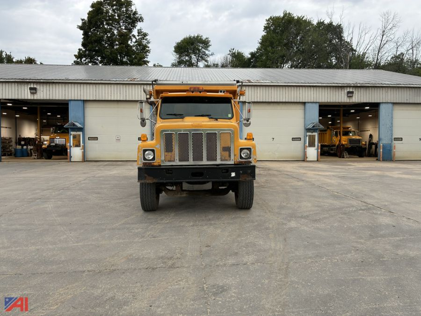 Dump truck front view showing large grille, rectangular headlights, and yellow cab with side mirrors; warehouse bays in background.