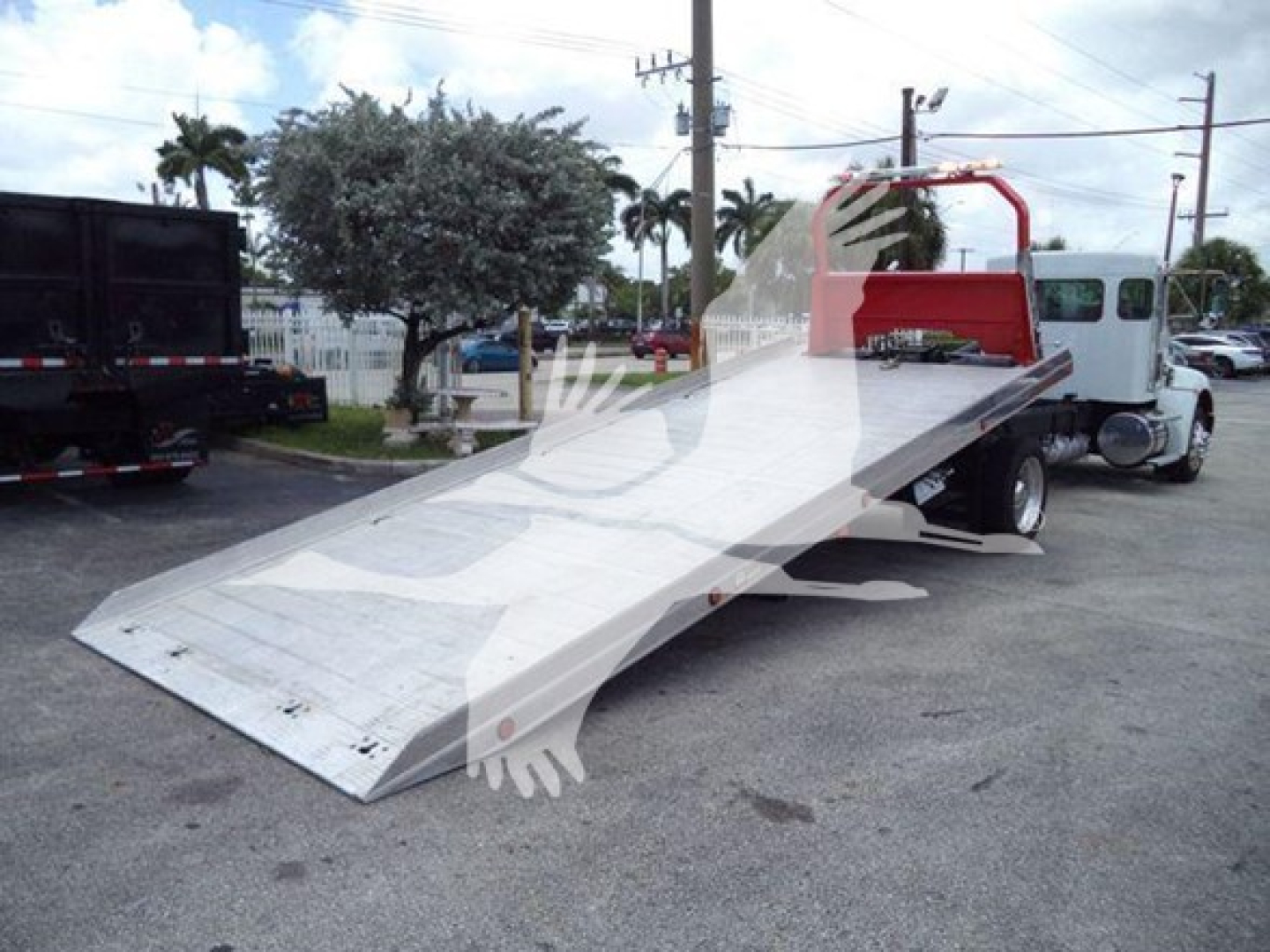 Kenworth rollback tow truck with a long flatbed and a red rollover rack at the back; no visible engine or dashboard; metal flatbed surface with slight wear; background shows other trucks and palm trees.