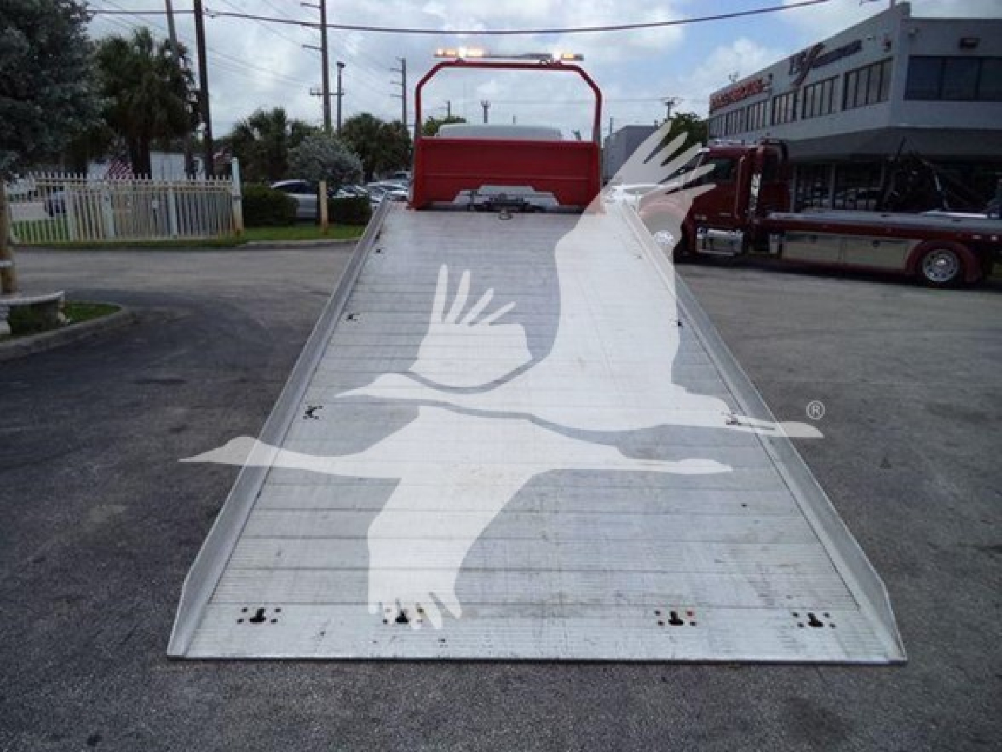 Kenworth T270 rollback: aluminum low-angle tilt bed with white bird logo watermark over the ramp; rear red cab area visible; business lot in background.