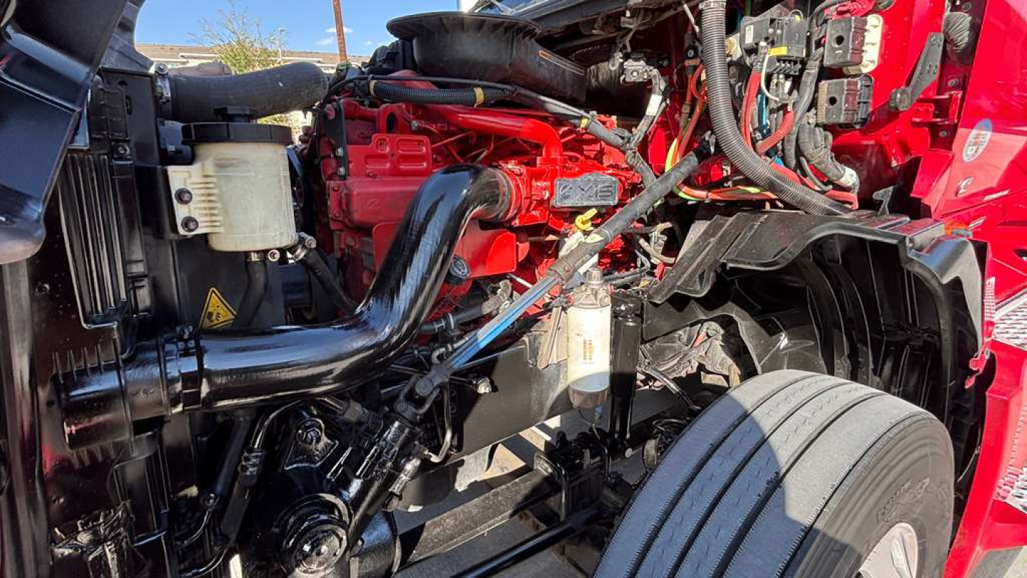 Truck engine bay of a 2022 International LT625 showing red engine components, black hoses, coolant reservoir, and front wheel with tire visible.