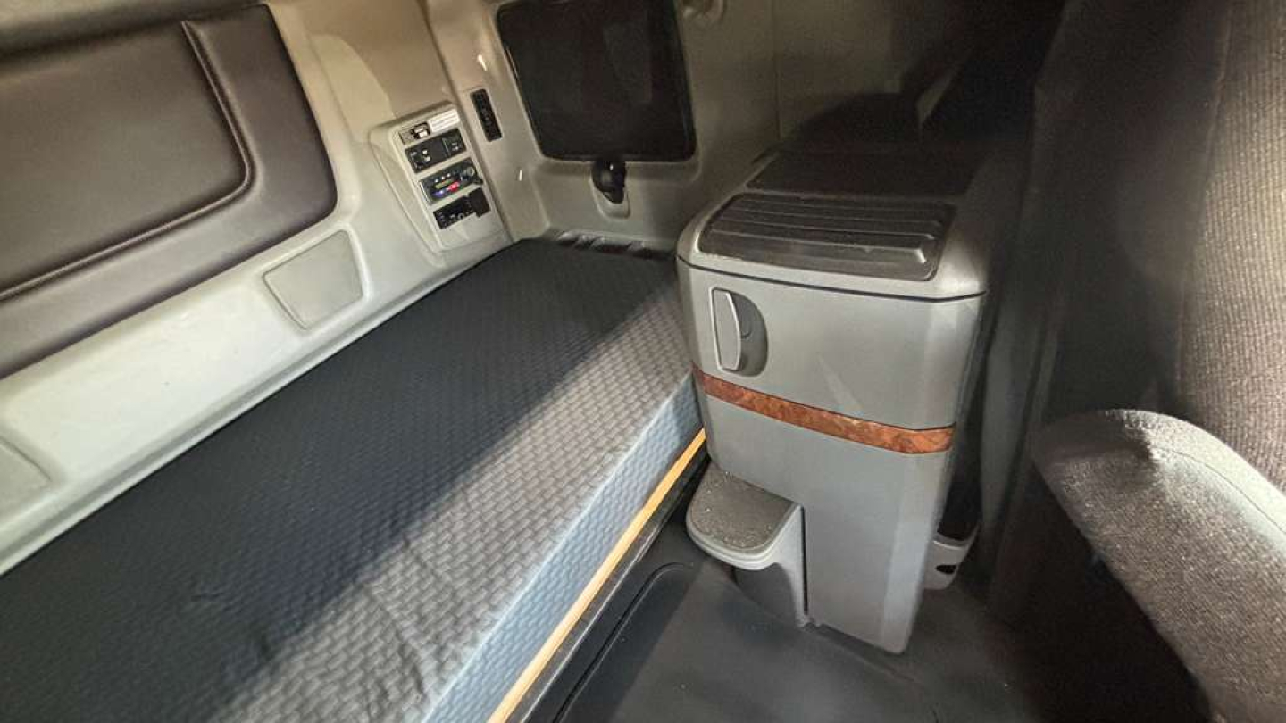 Truck cab interior showing sleeper berth with grey mattress, control panel, and storage unit with wood trim in a cab.