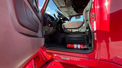 Truck cab interior with driver's seat, steering wheel, and door sill. Visible text includes a red fire extinguisher lying on the floor just under the dashboard and a small white label on the door frame.