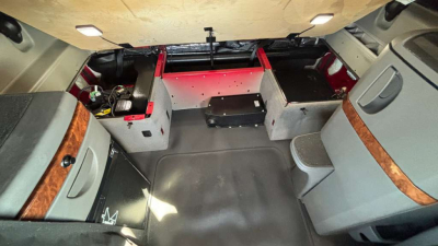 Truck cabin interior showing sleeper cab with storage compartments, grey seats with wood trim, and raised storage boxes behind the driver area; cargo area visible at rear.