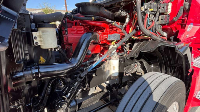 Truck engine bay of a 2022 International LT625 showing red engine components, black hoses, coolant reservoir, and front wheel with tire visible.