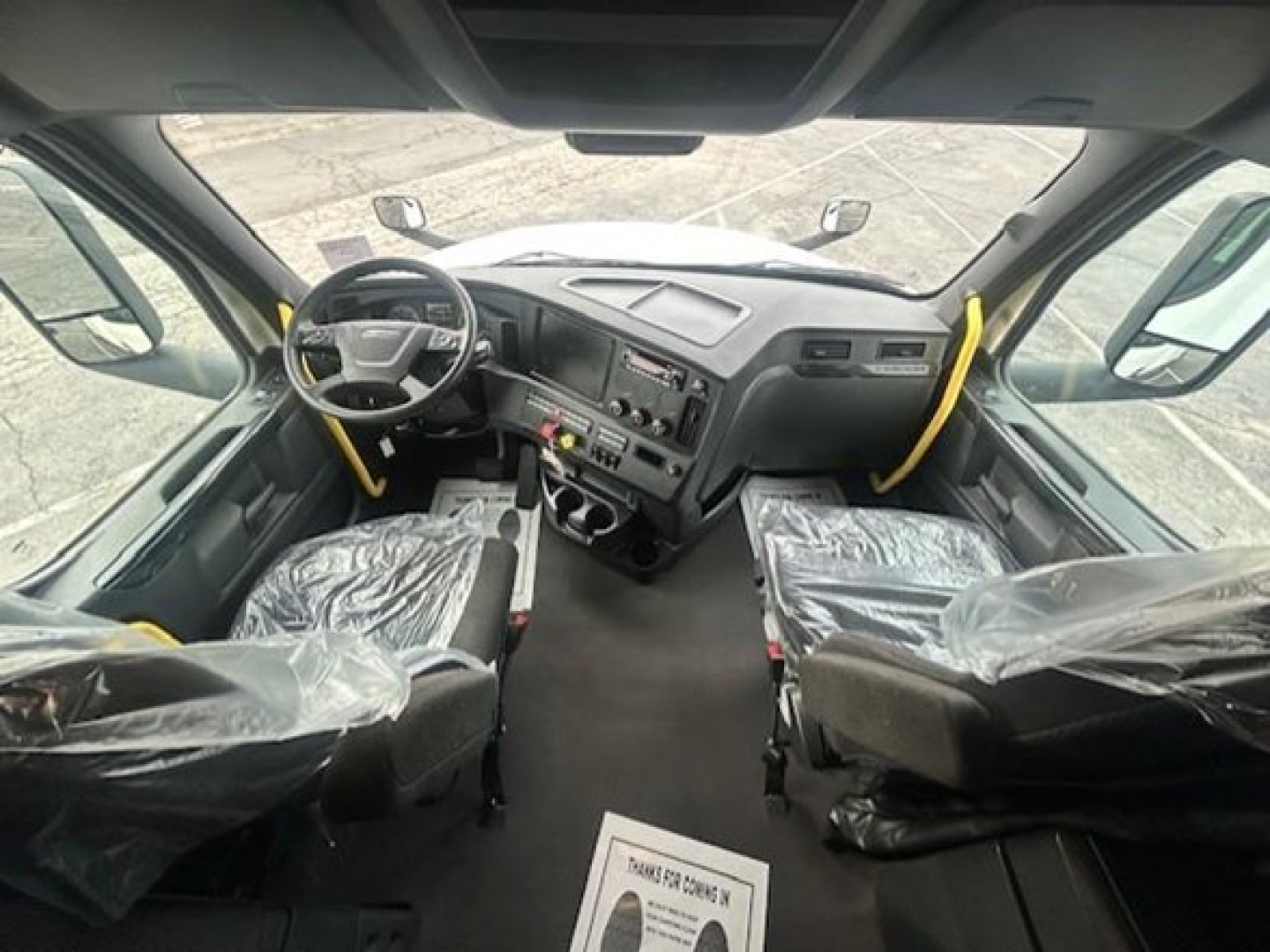 Cab interior of a 2024 Freightliner Cascadia 126 conventional sleeper showing steering wheel, dashboard, driver seat and passenger seat covered in plastic, with pedals and center console visible.