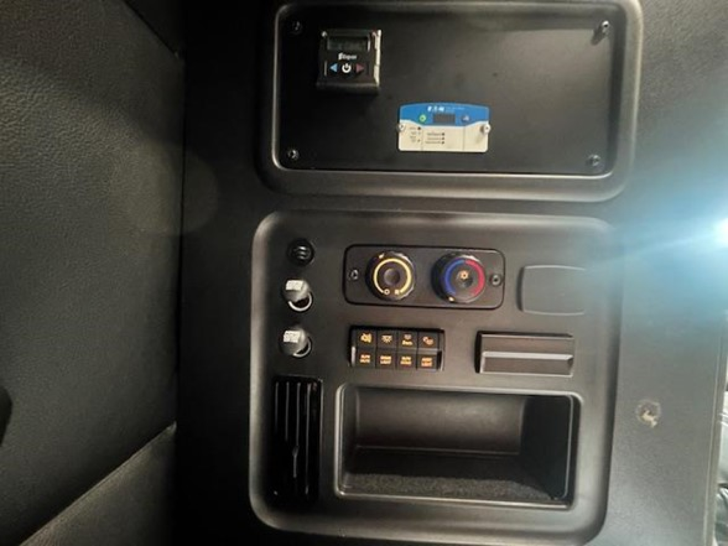 Truck cab interior dashboard with control panel and slots; visible labels include a blue and white sticker and a small black device in a recessed panel.