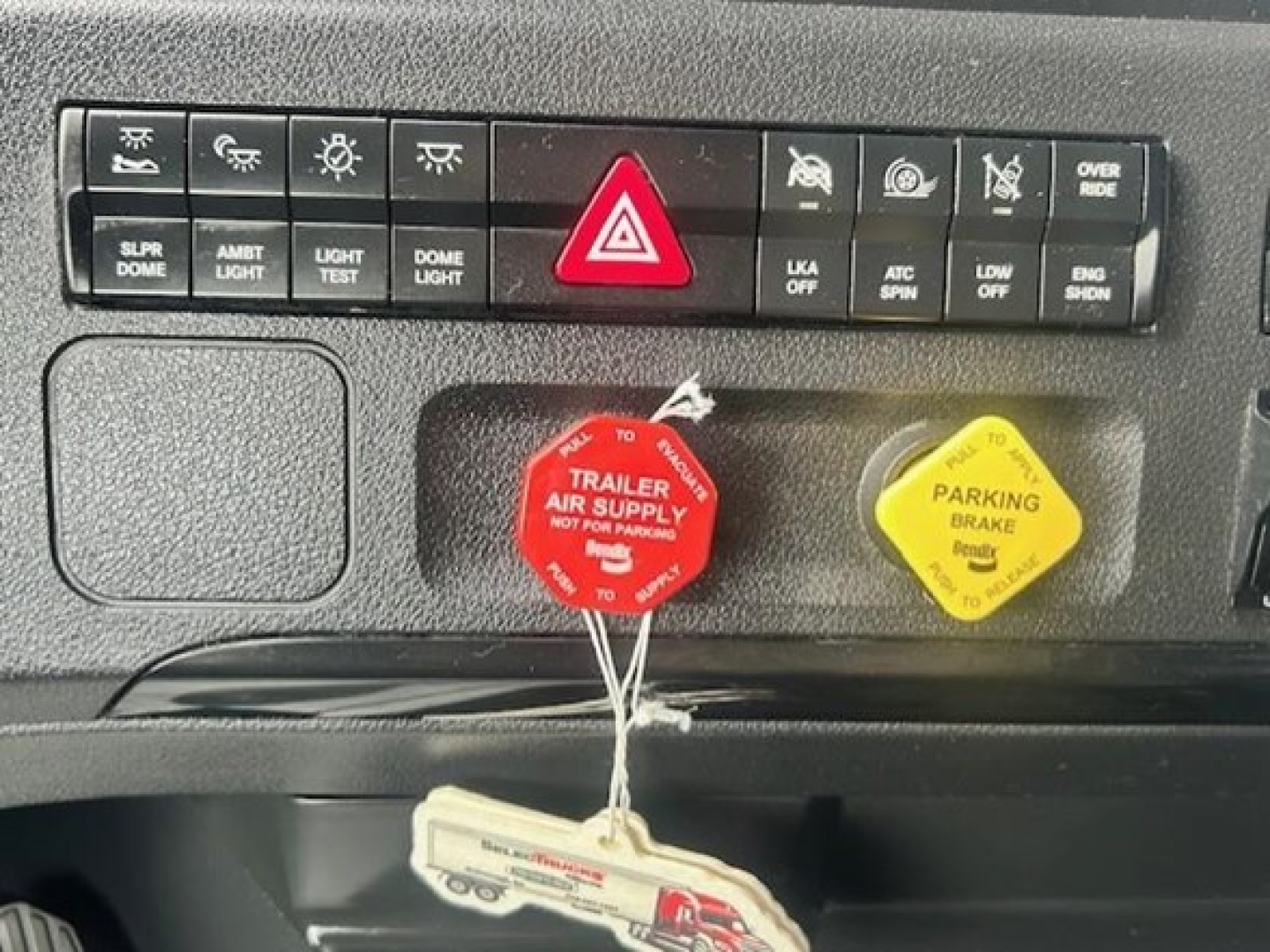 Freightliner Cascadia dash area showing hazard switch, red trailer air supply cap, yellow parking brake cap, and control buttons including lights and dome light.
