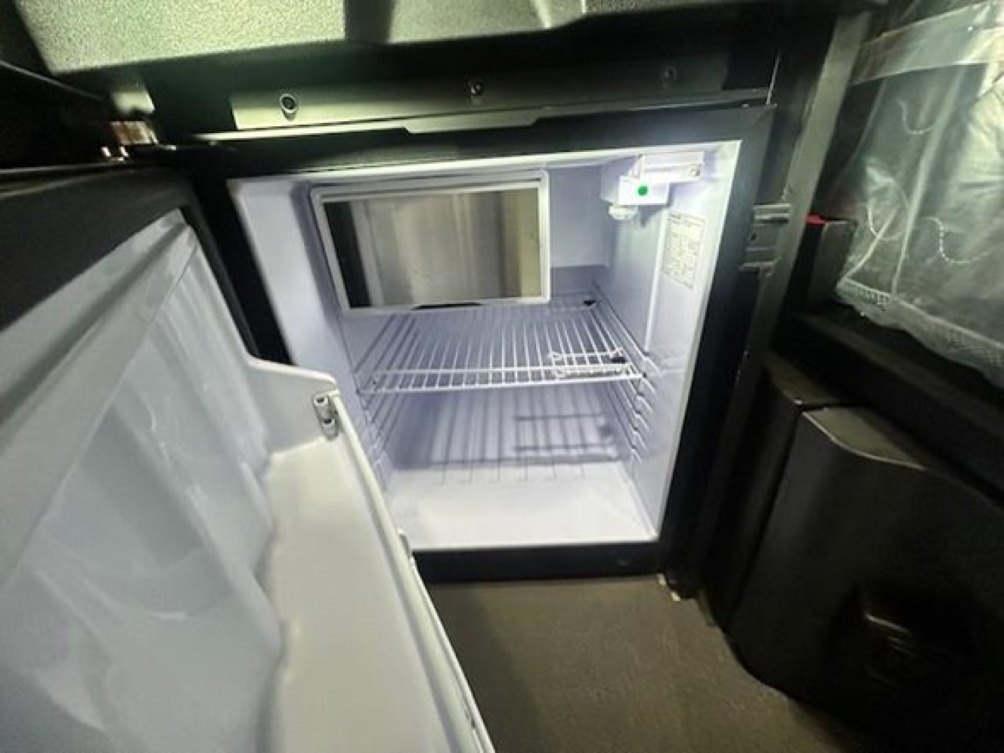 Refrigerator/freezer unit inside Freightliner Cascadia truck cab with open door and bare shelves.