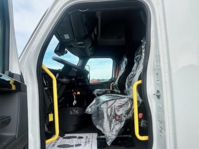 Truck cab interior of a 2024 Freightliner Cascadia 126 conventional sleeper showing driver seat wrapped in plastic and bright yellow entry handrails