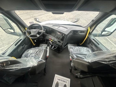 Cab interior of a 2024 Freightliner Cascadia 126 conventional sleeper showing steering wheel, dashboard, driver seat and passenger seat covered in plastic, with pedals and center console visible.
