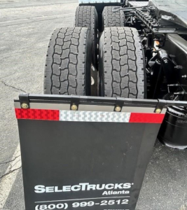 Rear view of a Freightliner Cascadia chassis showing dual rear axles and a black aluminum mudguard with reflective red-white striping. Text on mudguard reads SelectTrucks Atlanta with phone number partially visible.