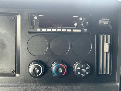 Dashboard area of a Freightliner Cascadia cab showing radio head unit, three circular gauges, climate control knobs, and power outlet.