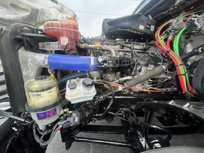 Truck engine bay of a Freightliner Cascadia showing coolant reservoir, power steering fluid reservoir, air intake hose, and various colored wiring harnesses.