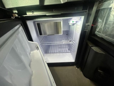 Refrigerator/freezer unit inside Freightliner Cascadia truck cab with open door and bare shelves.