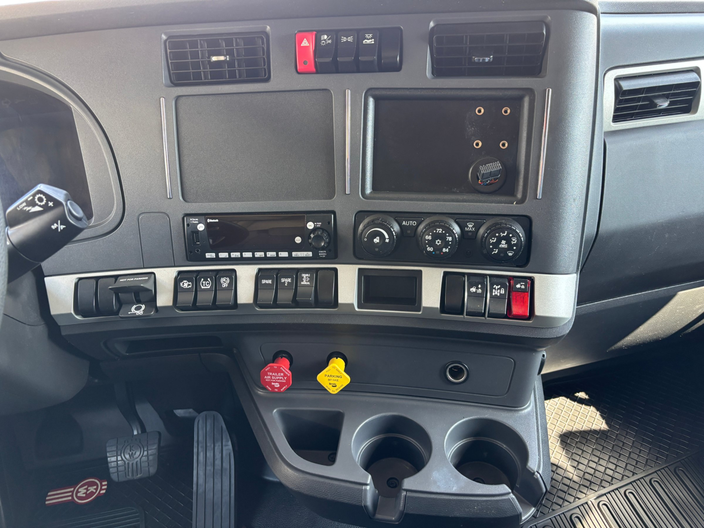 Dashboard of a Kenworth truck showing control switches, vents, and cup holders; visible text includes red and yellow caps and radio/display panels.