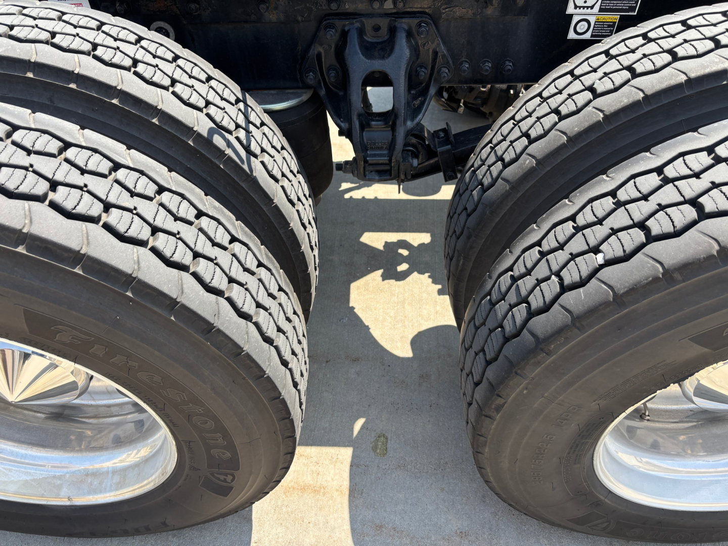 Kenworth T680 dual rear axles with dual tires and suspension components visible; view under trailer showing twin wheel assemblies and chassis cross member.