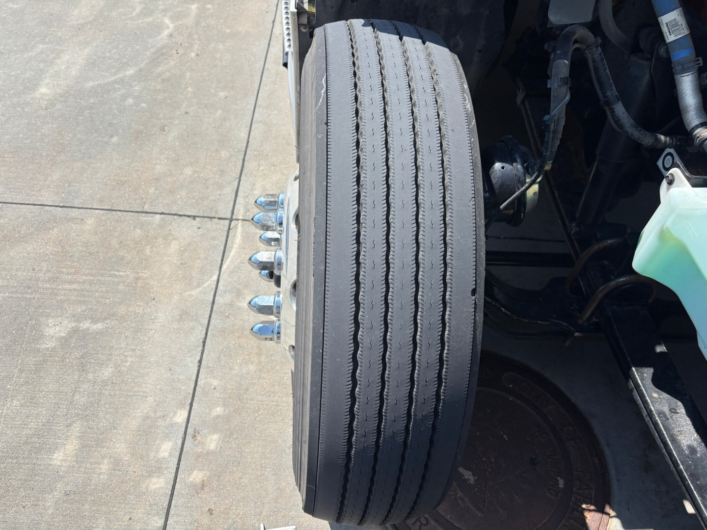 Truck front wheel and brake assembly showing wheel and lug nuts; portion of engine bay and reservoir visible.