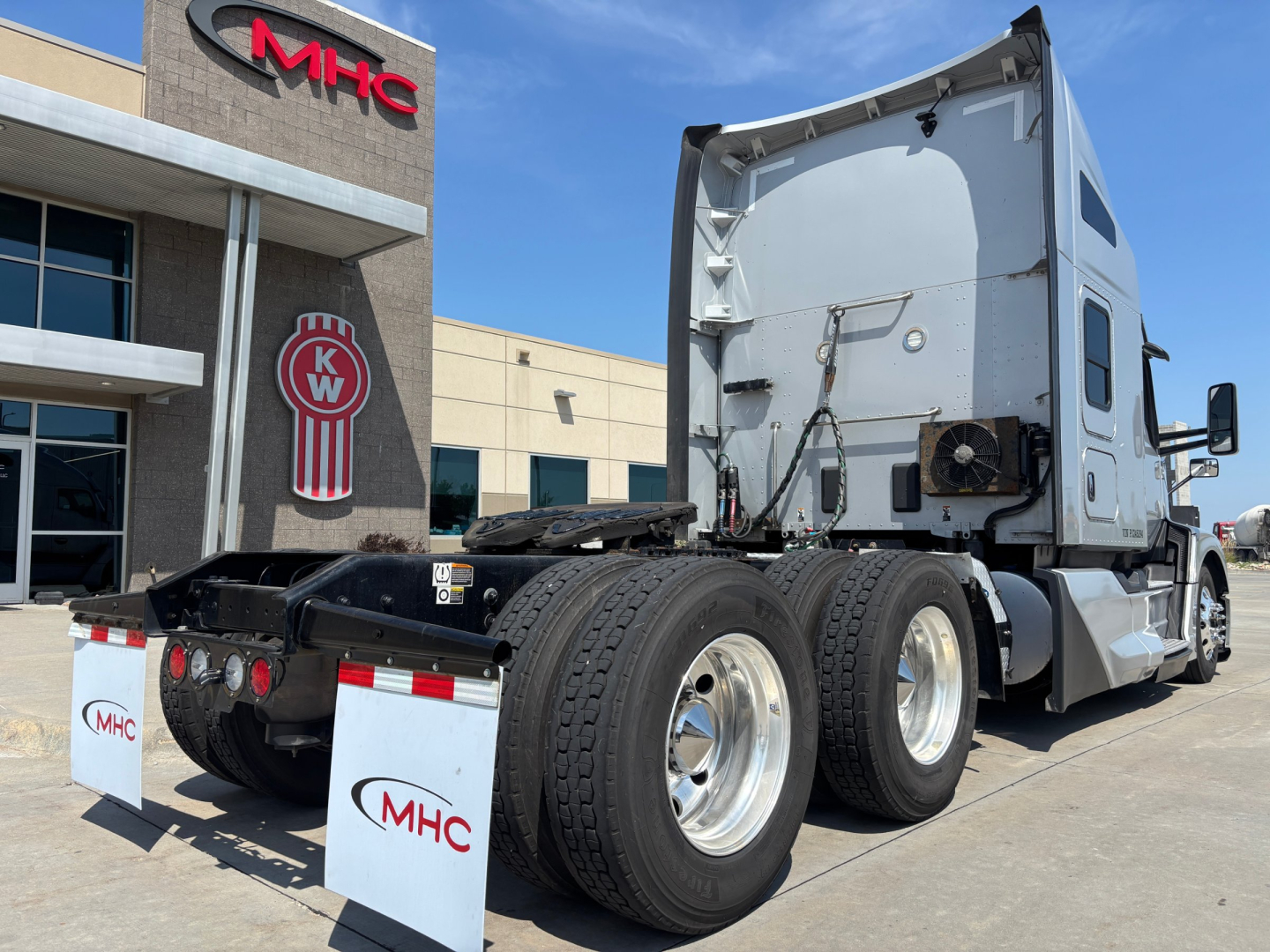 Kenworth T680 tractor in service with dual rear axles and sleeper cab, rear view showing tires and MHC logo on mudflaps.