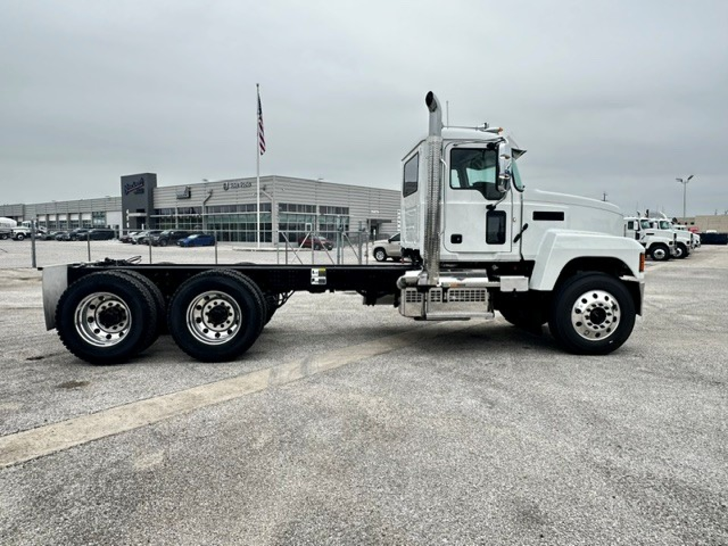 White Mack PI64 cab and chassis truck in a dealership lot, side view, showing empty flatbed chassis and dual rear axles with black wheels.