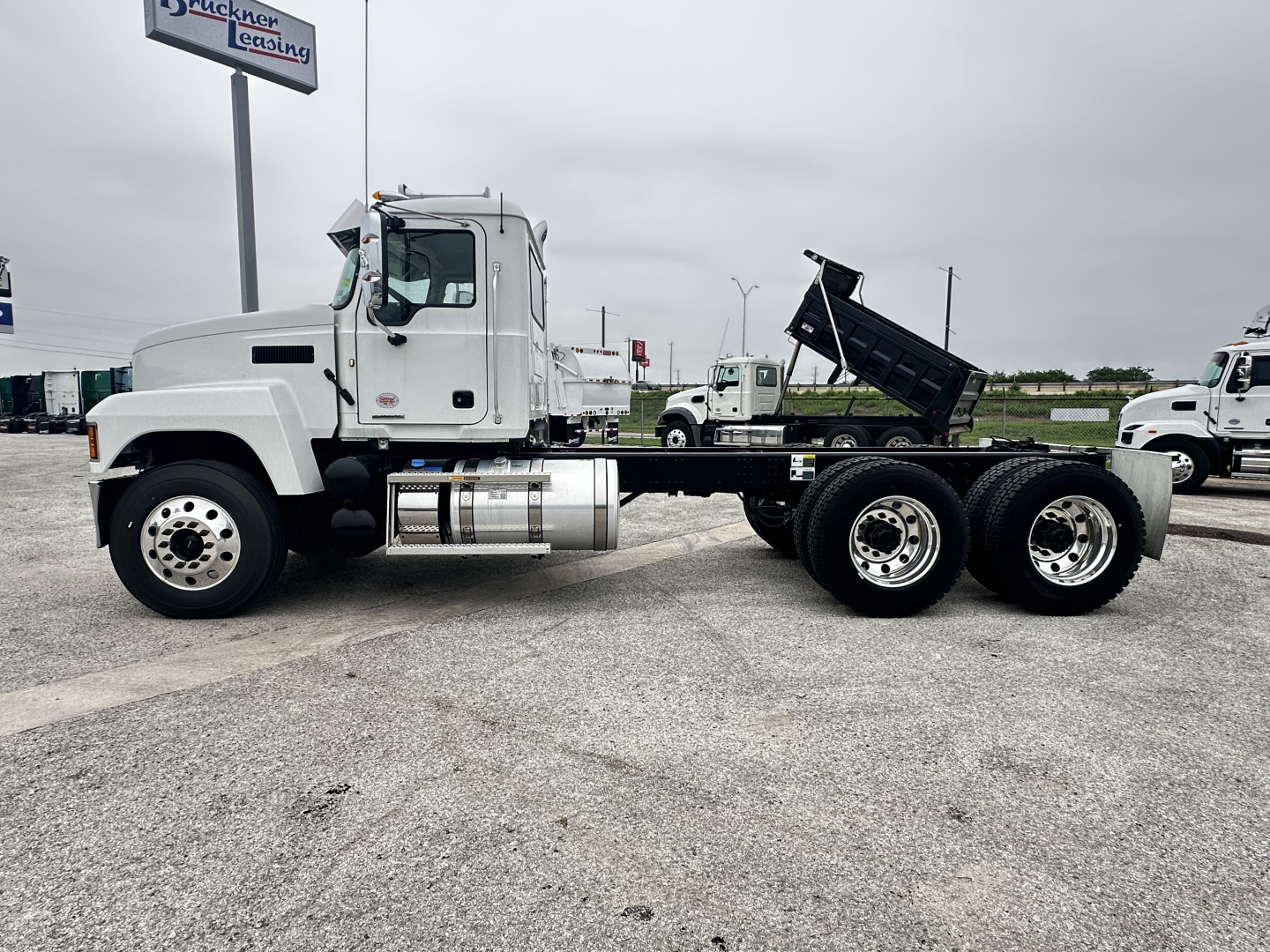 White Mack PI64 cab and chassis truck with dump bed partially visible, side view showing front cab, fuel tank, dual rear axles, and a raised dump bed in background.