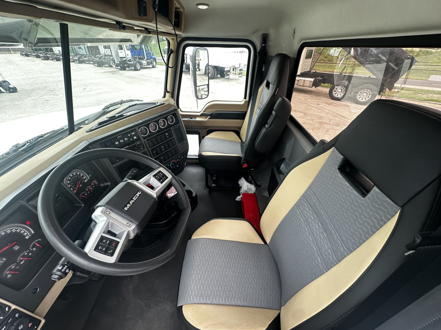 Cab interior of a 2026 Mack PI64 showing steering wheel, dashboard with gauges, grey and beige fabric seats, and entry to the sleeper area; view through front windshield and door window.