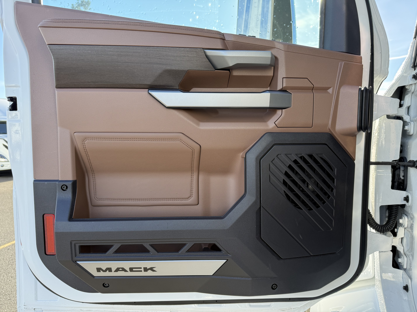 Interior of a 2026 Mack PR64T cabin door panel showing brown and gray trim, speaker grille, armrest, and door pocket with Mack branding.