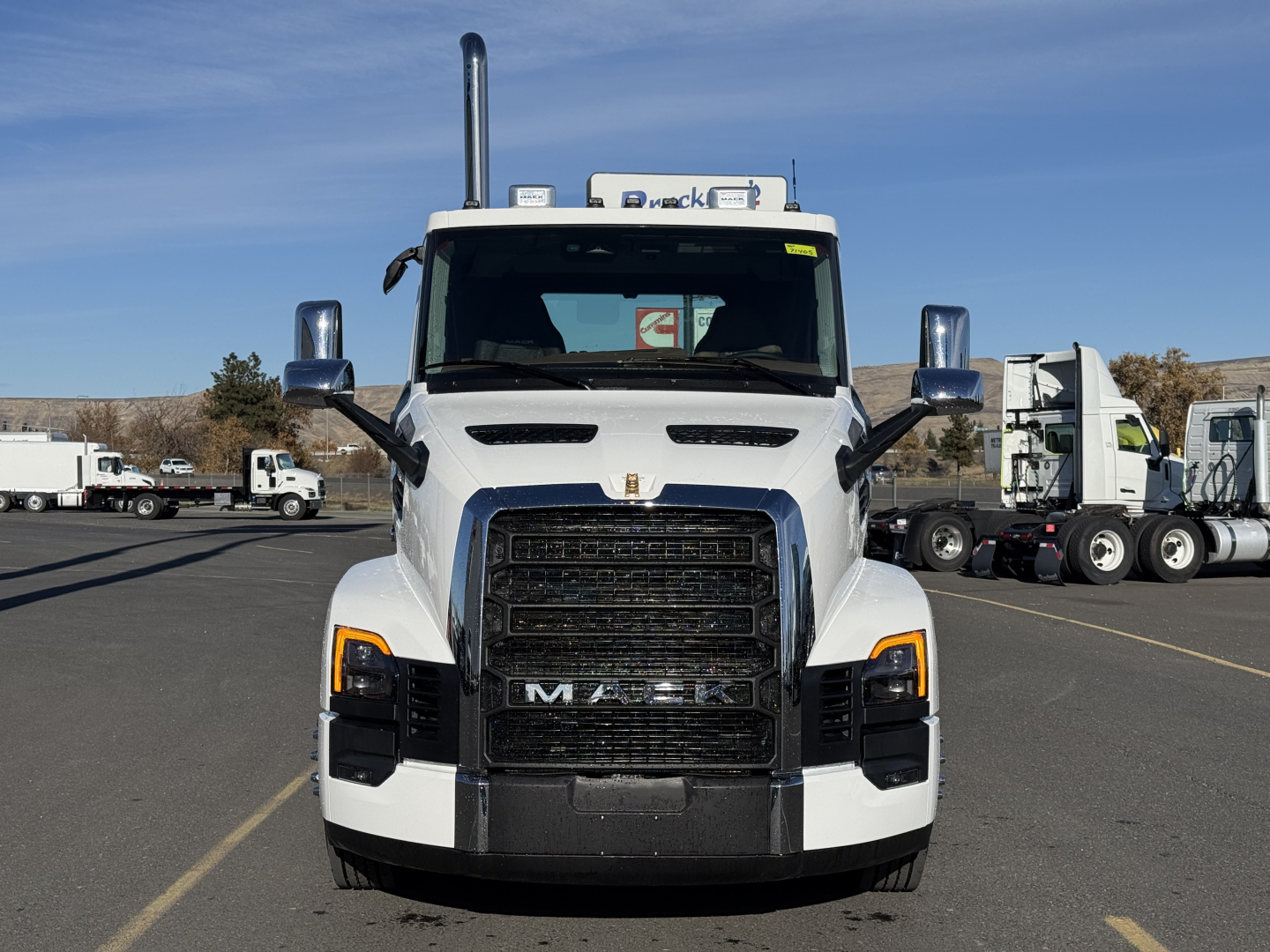 Mack PR64T truck front view showing large black grille, white cab, chrome mirrors, and stacked exhaust stacks with clear windshield and other trucks in background.
