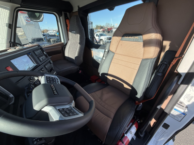 Truck interior: driver seat with Mack logo, steering wheel, dash controls, and large side window. Visible text on seat and control panels.