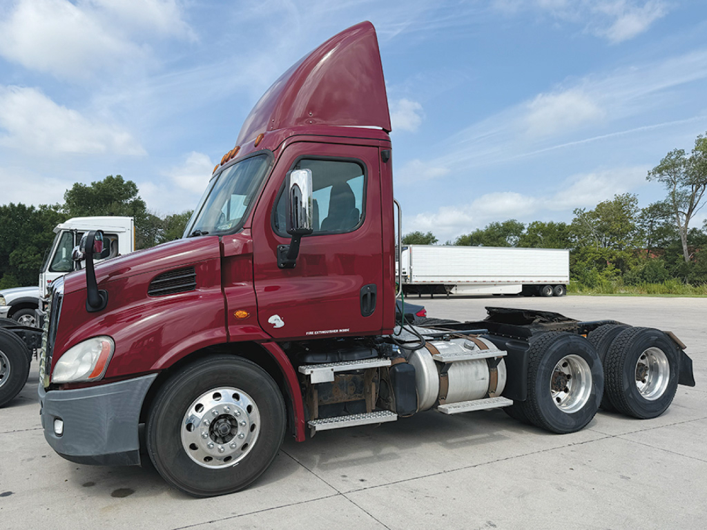 Truck side view of a maroon 2016 Freightliner Cascadia daytime cab tractor with twin rear axles, chrome fuel tanks, and a white trailer in the background.