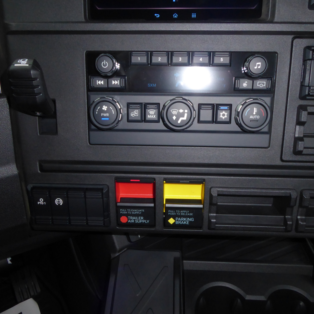 Dashboard controls of a 2026 Mack PR64T truck showing climate control knobs, radio panel, switch buttons, and two colored levers (red and yellow) for trailer air supply and parking brake.