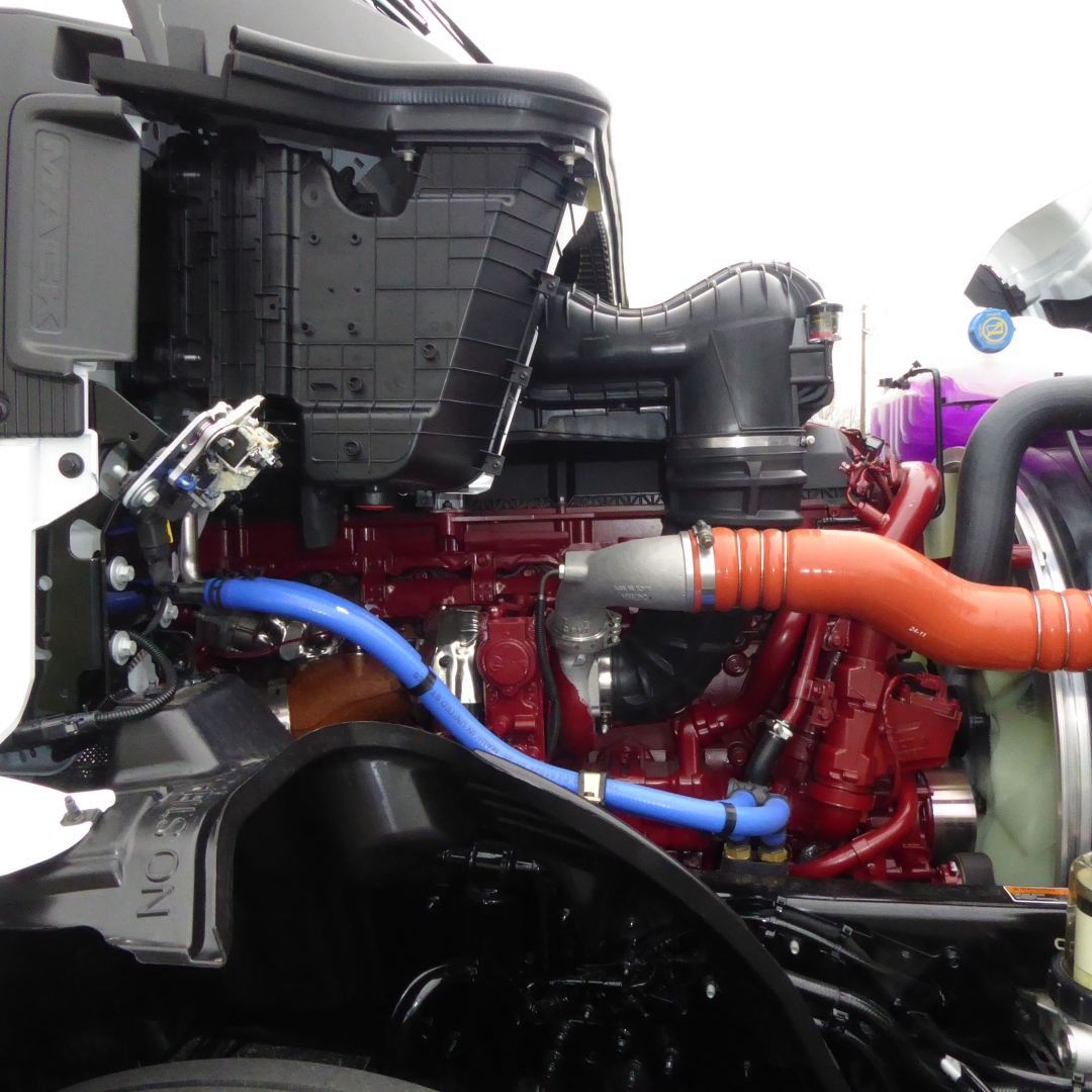 Mack PR64T engine bay showing red engine block, orange exhaust manifold, blue and black hoses, radiator and air intake components.