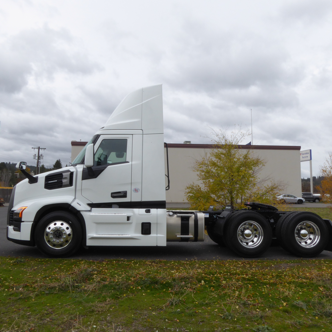 Truck chassis showing front cab and dual rear axles of a white 2026 Mack PR64T conventional daycab with chrome wheels.