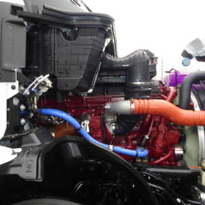 Mack PR64T engine bay showing red engine block, orange exhaust manifold, blue and black hoses, radiator and air intake components.
