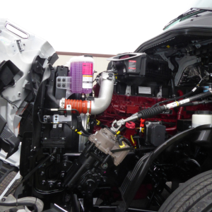 Engine bay of 2026 Mack PR64T showing red engine, pink coolant reservoir and various hoses and components.