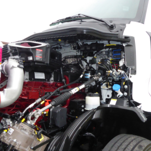 Engine bay of a 2026 Mack PR64T truck showing red engine, hoses, reservoir caps, and wiring in view from driver side.