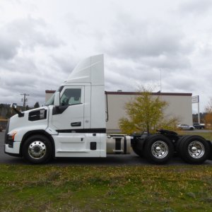 Truck chassis showing front cab and dual rear axles of a white 2026 Mack PR64T conventional daycab with chrome wheels.