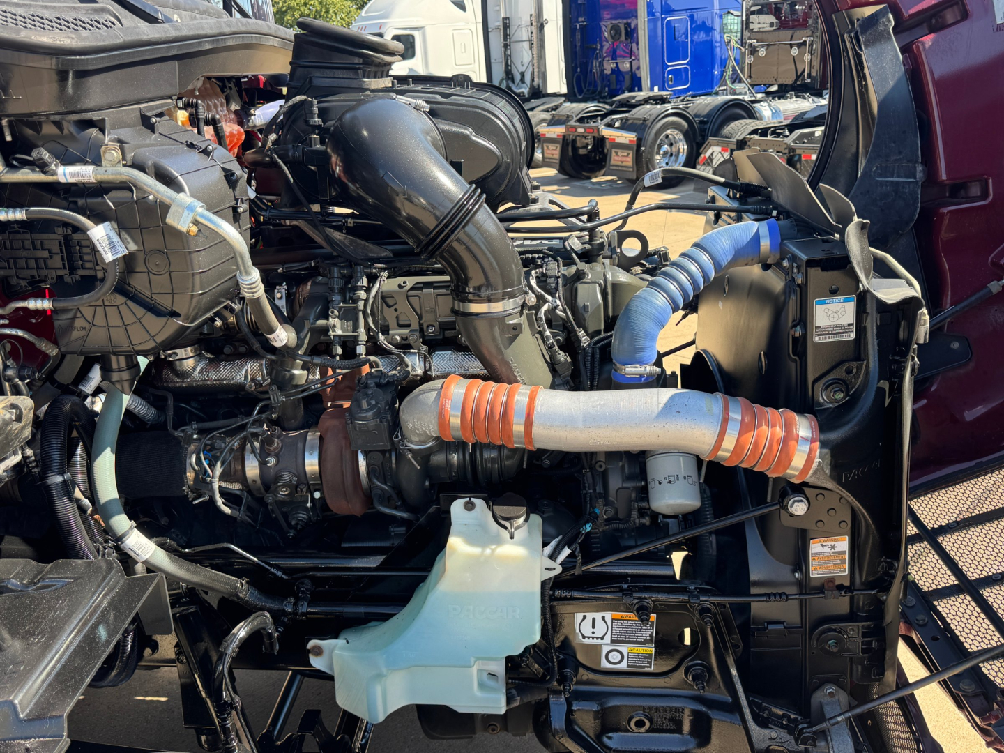 Truck engine bay with visible hoses, intercooler piping, and radiator on a 2023 Peterbilt 579. Engine components, blue and orange hoses, and intake tubing visible.