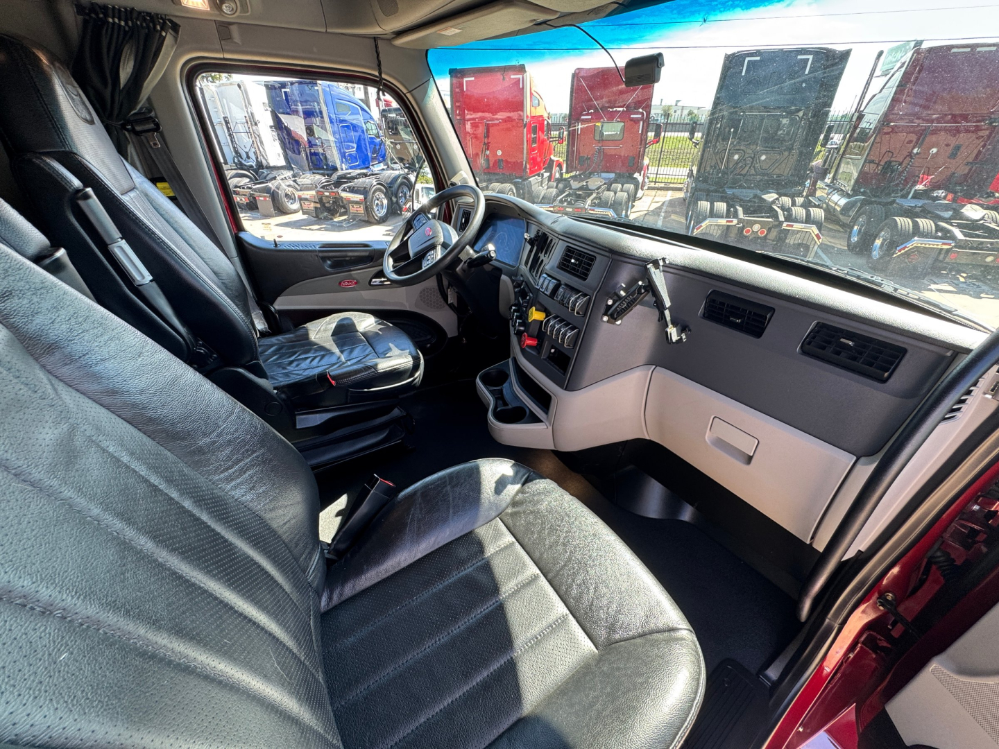 Peterbilt 579 truck cab interior showing steering wheel, dashboard with controls, driver's seat, and passenger seat; daylight outside with red trucks visible.