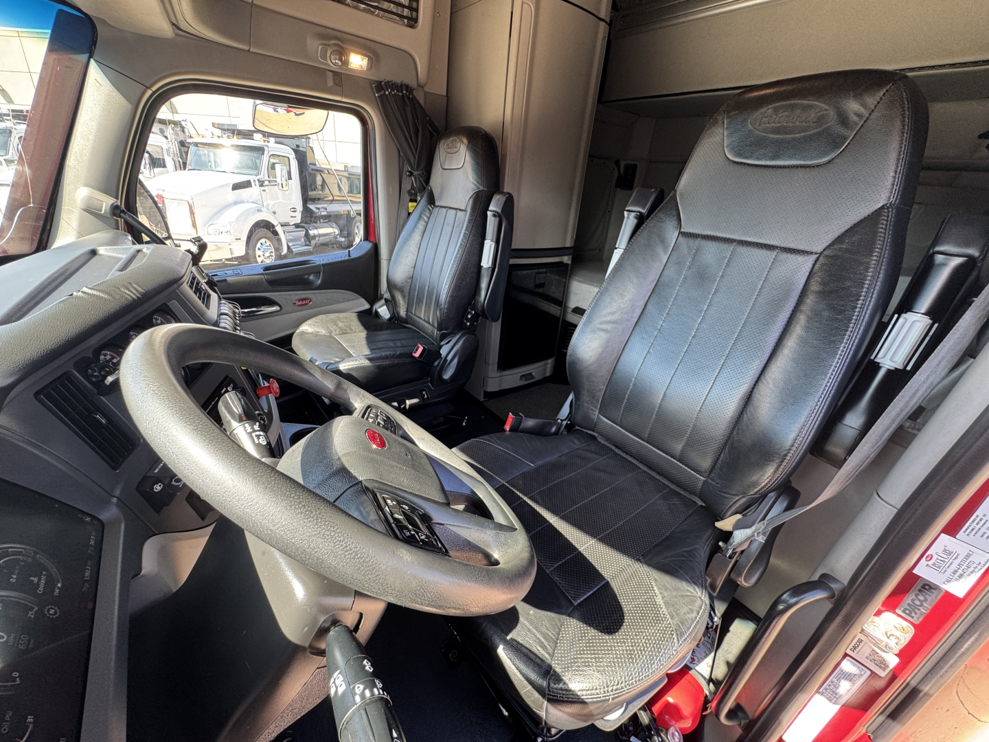 Truck cab interior showing steering wheel, driver seat, and passenger seat of a Peterbilt truck. Visible: dashboard controls, door, window, and seat belts.