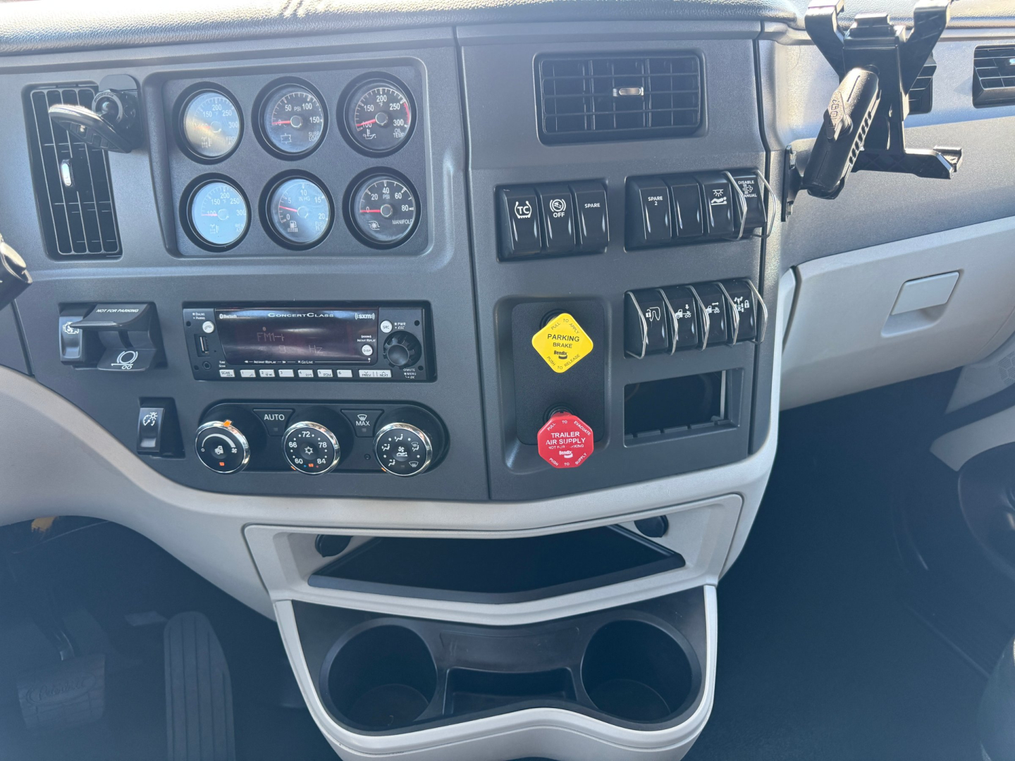 Peterbilt 579 dashboard view showing multiple gauges, switches, and a yellow parking brake badge; center stack includes radio, climate controls, and cup holders below.
