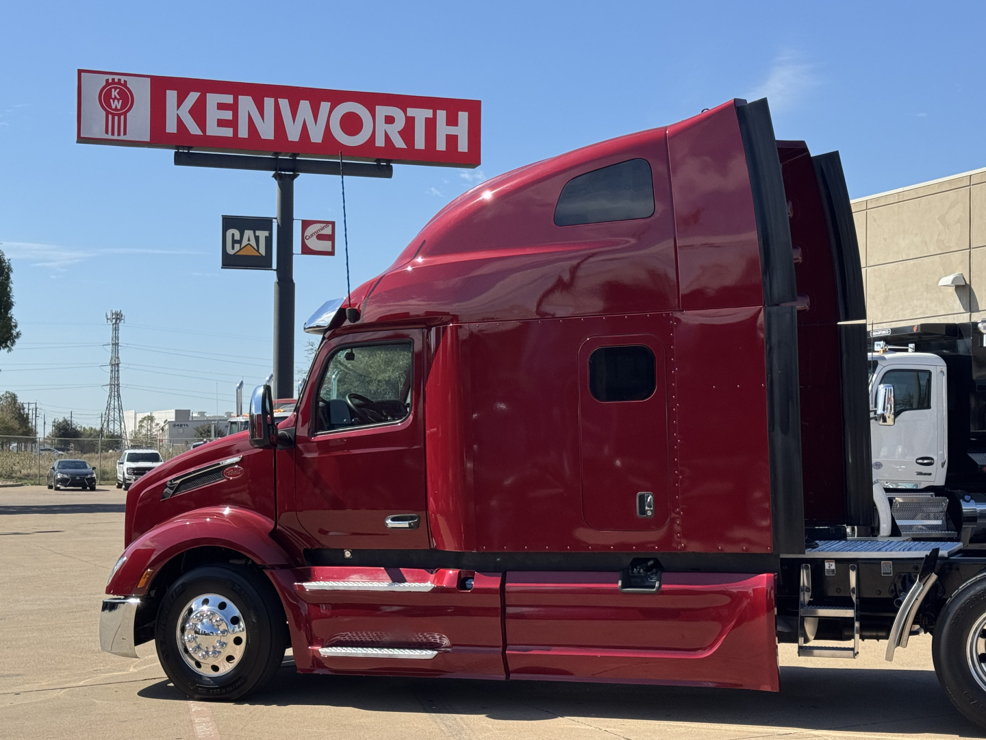 Peterbilt 579 tractor in red, side view showing cab and sleeper with Kenworth dealership sign in background.