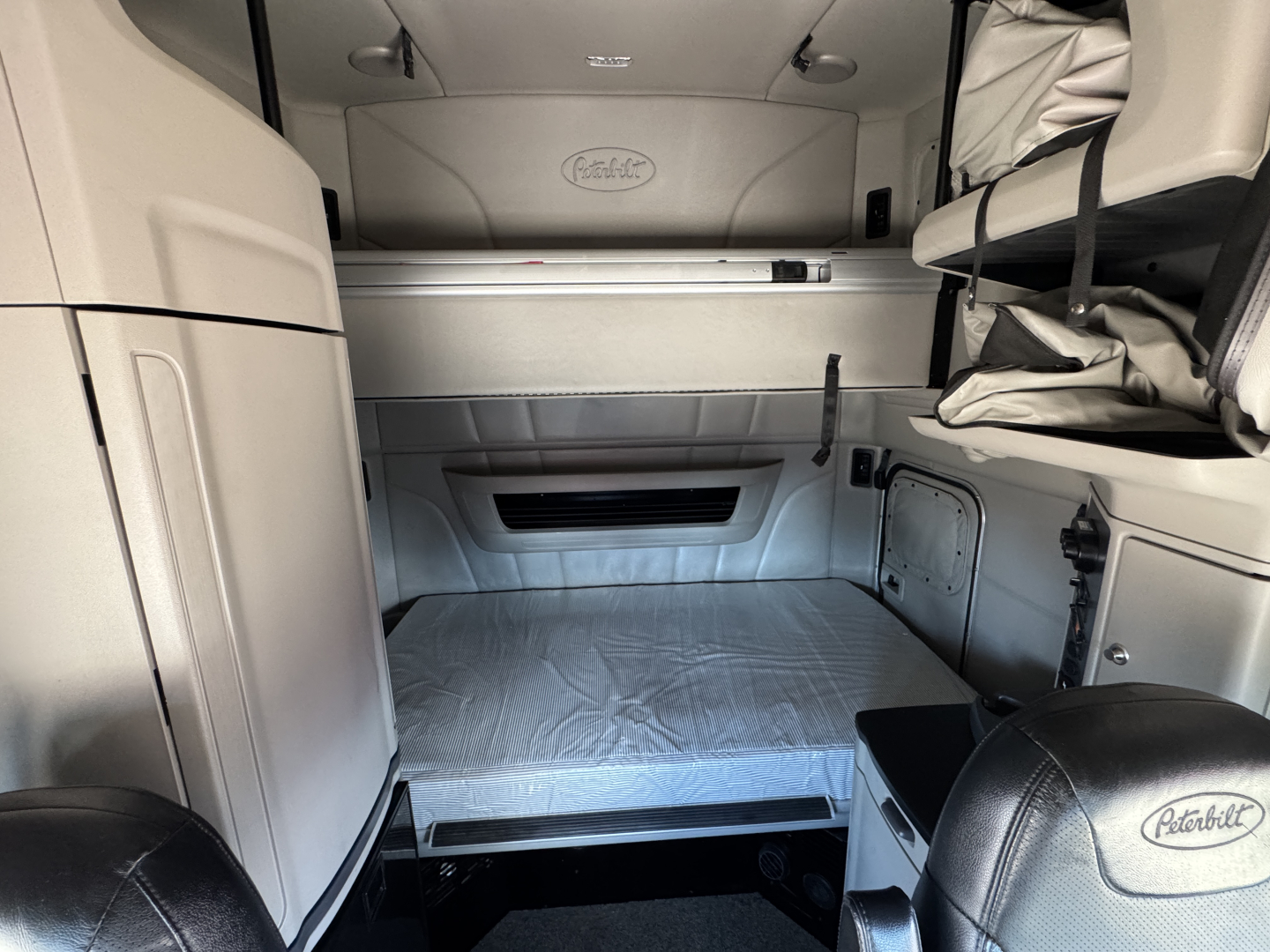 Peterbilt 579 cab interior showing sleeper bed area with overhead storage and controls, light gray interior, leather seats with Peterbilt logo.