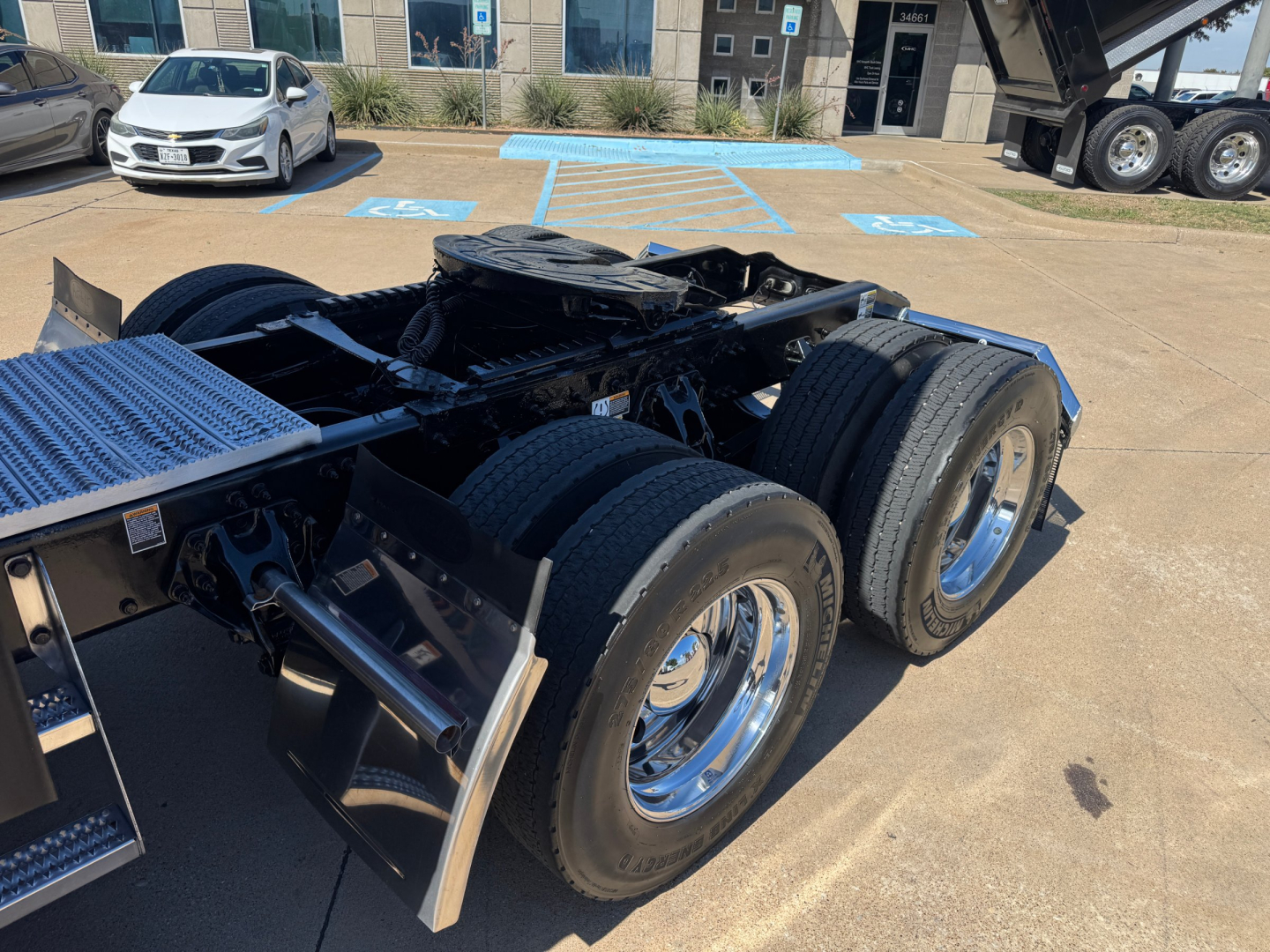 Peterbilt 579 chassis rear showing triple rear axle and dual tires, wheel rims, and fifth wheel area; background includes a dealership lot with a silver car and building.