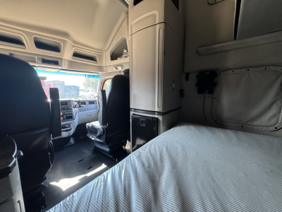Interior of a 2023 Peterbilt 579 sleeper cab showing driver's seat, passenger seat, dashboard, and elevated sleeper berth with bed and storage cabinet.