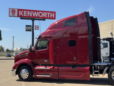 Peterbilt 579 tractor in red, side view showing cab and sleeper with Kenworth dealership sign in background.