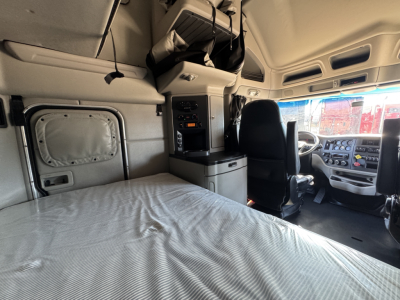 Truck cab interior of a 2023 Peterbilt 579 sleeper showing a bed, overhead storage, and driver seat with dashboard in view.