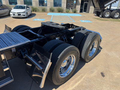 Peterbilt 579 chassis rear showing triple rear axle and dual tires, wheel rims, and fifth wheel area; background includes a dealership lot with a silver car and building.