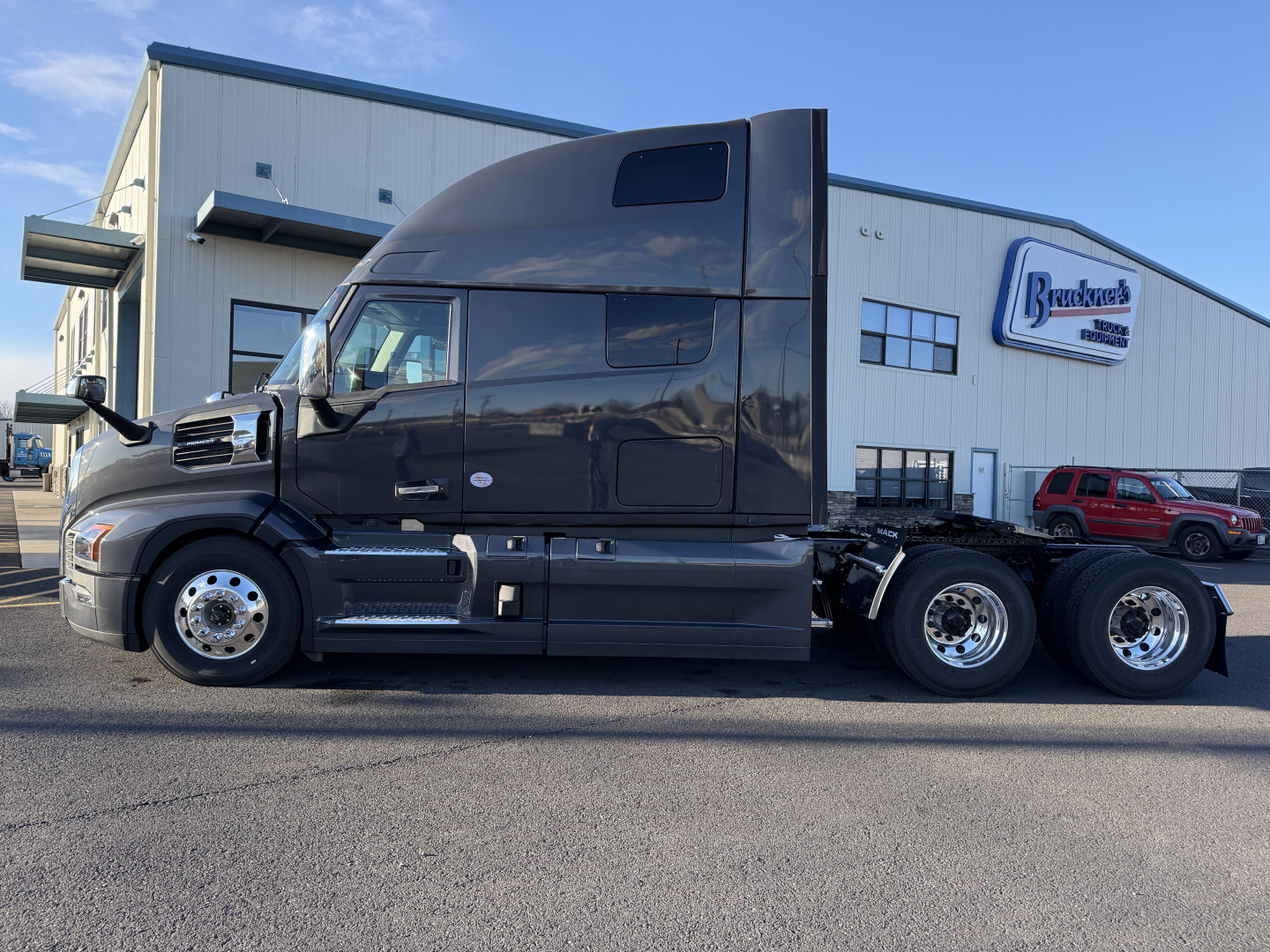 Truck side view of a black 2026 Mack PR64T76H conventional sleeper tractor, parked in front of a dealership building with a Brock’s logo sign. Visible text includes Mack, PR64T76H on listing, and wheel area.