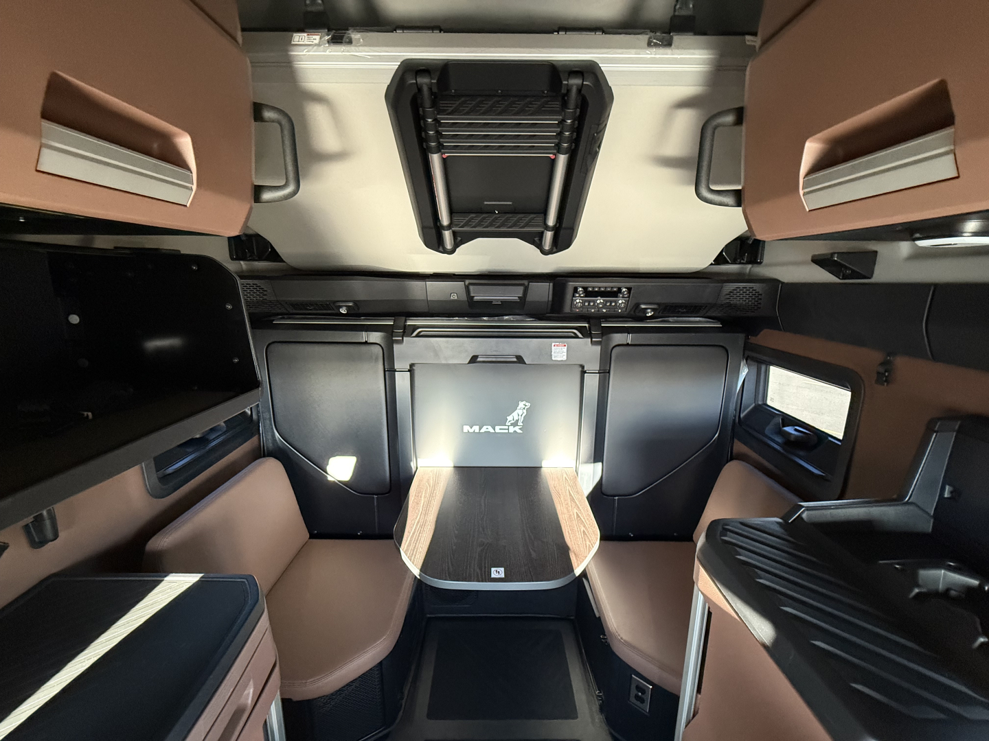Mack PR64T76H cab interior showing sleeper berth with tan upholstery, center console, overhead storage, and cockpit area.